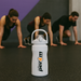 White Per4m water bottle in sharp focus on a gym floor with people performing HIIT training in the blurred background